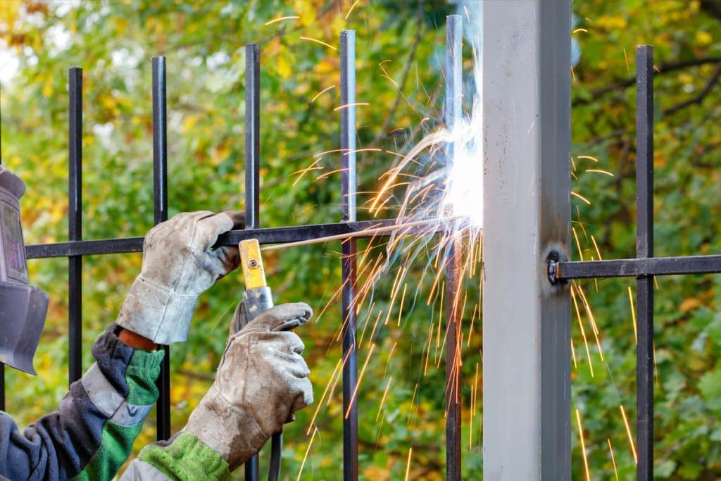 A worker wearing protective gloves welds a black metal fence to a gray steel post, creating bright sparks at the joint in an outdoor setting with green foliage in the background.