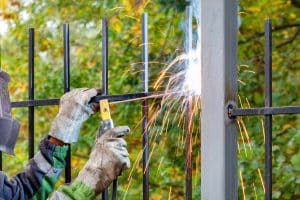 A worker wearing protective gloves welds a black metal fence to a gray steel post, creating bright sparks at the joint in an outdoor setting with green foliage in the background.